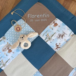 Könnte beinhalten: Eine Babydecke mit Patchwork-Design in Blau, Beige und Braun. Die Decke hat eine weiche Hasenrassel und ein Etikett mit einem Herz. Der Name "Florentin" und das Datum "05. Juni 2025" sind aufgestickt.