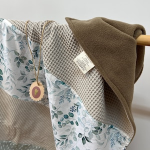 Pode incluir: Um cobertor de bebé em tecido waffle bege com um estampado floral verde e branco de um lado. O cobertor tem um forro de lã marrom do outro lado. O cobertor tem uma etiqueta que diz "EXCEED".