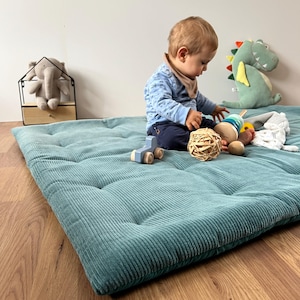 Puede incluir: Una alfombra de juego de pana azul verdosa con un bebé sentado en ella jugando con juguetes. La alfombra tiene un diseño acolchado y es perfecta para una sala de juegos o una guardería.