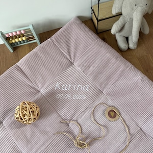 May include: A dusky pink corduroy baby blanket with a patchwork design. The name "Karina" and the date "02.05.2020" are embroidered in white. A small wooden tag and a woven ball are also present.