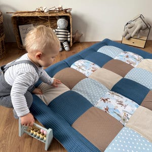 Op de afbeelding: Een patchwork speelmat in blauw, beige en bruin, met een getextureerde rand. De mat heeft verschillende patronen, waaronder stippen en vliegtuigontwerpen. Een kind speelt met een telraam op de houten vloer.
