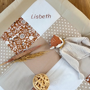 Könnte beinhalten: Eine Patchwork-Babyspielmatte mit dem Namen "Lisbeth" in Burgunderrot gestickt. Die Matte weist verschiedene Stoffe auf, darunter Blumen-, Tupfen- und Streifenmuster in Braun, Beige und Weiß. Ein Stofftier-Fuchs und dekorative Trockenpflanzen sind ebenfalls vorhanden.