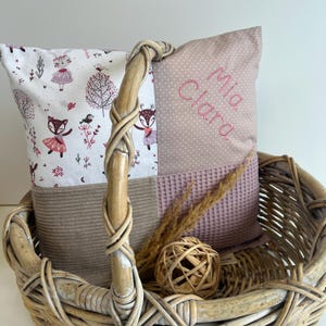 Op de afbeelding: Een roze en bruin patchwork kussen met de naam "Mia Clara" geborduurd erop. Het kussen ligt in een rieten mand.