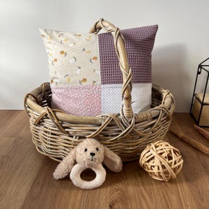 Op de afbeelding: Een rieten mand met een handvat, gevuld met een patchwork kussen met een bloemenpatroon. Een bruin knuffeldier met een ring ligt voor de mand, en een geweven bal ligt rechts.