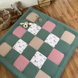 May include: A green quilted play mat with a patchwork design. The patchwork includes squares of pink, beige, and patterned fabric with forest animal illustrations. A wooden toy and stuffed animal are on the mat.