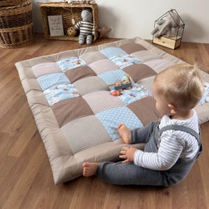 Könnte beinhalten: Eine beige Spielmatte mit Patchwork-Design aus blauen, braunen und gemusterten Quadraten. Ein Baby in grauen Latzhosen sitzt auf der Matte, mit einem Holzspielzeug. Die Matte ist für Babyspiele und Bodenaktivitäten konzipiert.