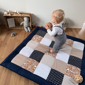 Könnte beinhalten: Eine Patchwork-Spielmatte mit marineblauem Rand. Die Matte hat Quadrate aus verschiedenen Stoffen, darunter Punkte, Blumenmuster und Streifen in neutralen Tönen. Ein Baby in grauen Latzhosen steht auf der Matte und hält einen Ball.