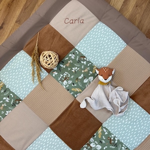 May include: A multi-coloured patchwork play mat with a brown border. The mat is made of different fabrics with various patterns, including a green fabric with animal prints, a brown fabric with a corduroy texture, and a light blue fabric with white dots. The mat has the name "Carla" embroidered in the top centre square.