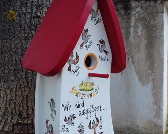 May include: A white birdhouse with a red roof and a yellow sign that says "SPATZENNEST 2019". The birdhouse is decorated with painted birds and the names of children: Edda, Bente, Moritz, Nylla, Klis, Pius, Luis, Conner, and Simone. The text on the birdhouse says "Wir sind ausgeflogen..."