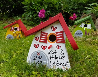 May include: Three colorful birdhouses with red, white, and green paint. The birdhouses have heart designs and the text "Julia + Romeo", "Kati & Holgers Liebesnest", and "T&A" on them. The birdhouses are sitting on a bed of green grass.