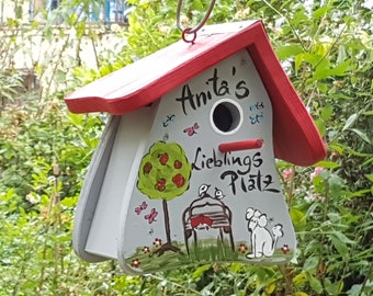 May include: A gray and red birdhouse with a red roof and a painted scene of a tree, birds, and a dog. The birdhouse has a hole for birds to enter and the text "Anita's Lieblings Platz" is painted on the front.
