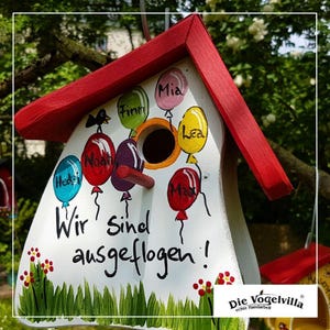 Könnte beinhalten: Ein weißes Vogelhaus mit rotem Dach, verziert mit bunten Luftballons und den Namen Hedi, Noah, Finn, Mia, Lea und Max. Der deutsche Text "Wir sind ausgeflogen!" ist auf der Vorderseite geschrieben. Das Vogelhaus hat ein rundes Einflugloch und ist von gemalten Blumen und Gras umgeben.