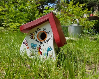 Puede incluir: Una casita de pájaros blanca con un techo rojo y un agujero redondo para que los pájaros entren. La casita está pintada con mariposas de colores y el texto "Wir sind ausgeflogen!" que se traduce como "¡Hemos volado!" en español. La casita está sentada en un lecho de hierba verde.