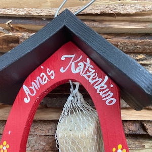 May include: A red wooden bird feeder with a black roof. The feeder has a white inscription that reads "Anna's Katzenkino". A white suet cake is hanging from the feeder in a mesh bag.