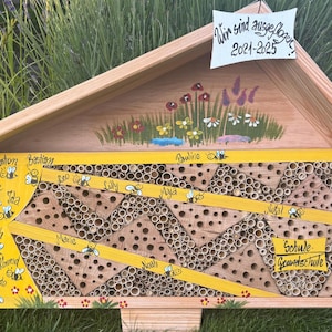 Könnte beinhalten: Ein hölzernes Bienenhaus mit Satteldach, bemalt mit Blumen und dem Text "Wir sind ausgeflogen 2021-2025". Die Vorderseite hat eine gelbe Tafel mit Namen und Bienenillustrationen sowie einen Abschnitt mit Bambusröhren.