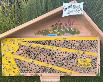 May include: A wooden bee house with a pitched roof, painted with flowers and the text "Wir sind ausgeflogen 2021-2025". The front features a yellow panel with names and bee illustrations, and a section with bamboo tubes.