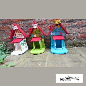 May include: Three brightly painted, handcrafted birdhouses shaped like boots. Each has a red roof, a small porch, and is painted in white, green, and blue. The birdhouses feature German text and are decorated with colourful polka dots.