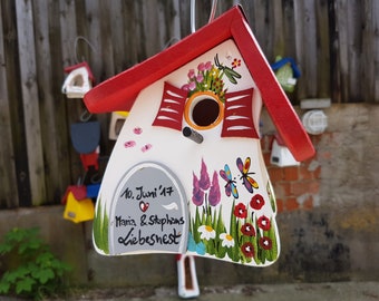 Puede incluir: Una casita de pájaros blanca con un techo rojo y una placa gris que dice "10. Juni '17 Maria & Stephens Liebesnest". La casita está decorada con flores, mariposas y una libélula.
