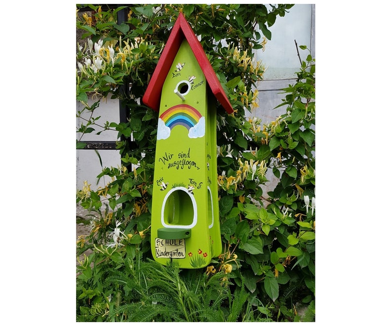 Op de afbeelding: Een groen en rood vogelhuisje met een regenboog die op de voorkant is geschilderd. Het vogelhuisje heeft een kleine opening waar vogels naar binnen kunnen en een bord met de tekst "Schule Kindergarten".