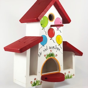 Könnte beinhalten: Ein weißes Vogelhaus mit rotem Dach und einem bemalten Design aus bunten Luftballons. Der Text auf dem Vogelhaus lautet "Wir sind ausgeflogen..."