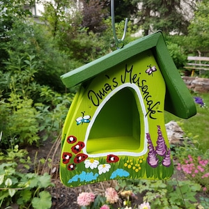 May include: A green birdhouse with a white painted opening and the words "Oma's Meisencafe" painted on the front. The birdhouse is decorated with painted flowers, butterflies, and ladybugs.