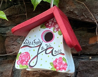 May include: A white birdhouse with a red roof, painted with pink flowers and green leaves. The birdhouse has a small hole for birds to enter and the text "Liz & Frank Ja 26. Juli 2022" is written on the front.