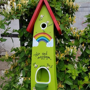 Op de afbeelding: Een groen houten vogelhuisje met een rood dak. Het vogelhuisje heeft een regenboog geschilderd op de voorkant en de woorden "Wir sind ausgeflogen..."  Het vogelhuisje heeft ook de namen "Dan", "Connor", "Eric" en "Tom G." erop geschilderd. Het vogelhuisje staat voor een groene struik met witte bloemen.