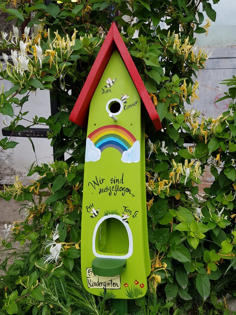 Op de afbeelding: Een groen houten vogelhuisje met een rood dak. Het vogelhuisje heeft een regenboog geschilderd op de voorkant en de woorden "Wir sind ausgeflogen..."  Het vogelhuisje heeft ook de namen "Dan", "Connor", "Eric" en "Tom G." erop geschilderd. Het vogelhuisje staat voor een groene struik met witte bloemen.