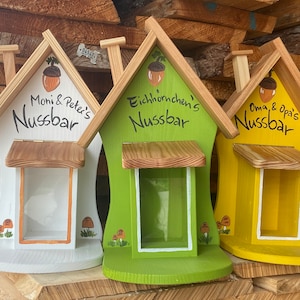 Puede incluir: Tres comederos de pájaros de madera con forma de casas con una pequeña abertura para que los pájaros puedan acceder a la comida. Las casas están pintadas en diferentes colores: blanco, verde y amarillo. Cada casa tiene un letrero con un nombre y la palabra "Nussbar", que significa "barra de nueces" en alemán. Los letreros dicen "Moni & Peter's Nussbar", "Eichhörnchen's Nussbar" y " Oma & Opa's Nussbar".