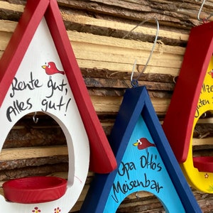 May include: Three wooden bird feeders painted in red, blue, and yellow with a red bird illustration on each. The feeders have a small red, yellow, and blue tray at the bottom. The feeders have handwritten messages on them: "Rente gut, alles gut!" on the red feeder, "Opa Ottos Meisenbar" on the blue feeder, and "Allzeit guten Flug!" on the yellow feeder.