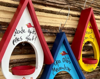 Pode incluir: Três comedouros de pássaros de madeira pintados de vermelho, azul e amarelo com uma ilustração de um pássaro vermelho em cada um. Os comedouros têm uma pequena bandeja vermelha, amarela e azul na parte inferior. Os comedouros têm mensagens escritas à mão: "Rente gut, alles gut!" no comedouro vermelho, "Opa Ottos Meisenbar" no comedouro azul e "Allzeit guten Flug!" no comedouro amarelo.