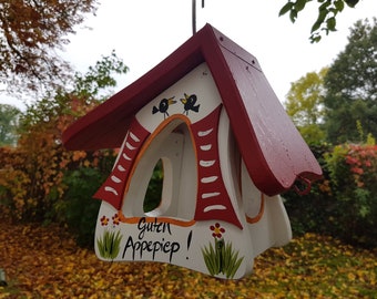 May include: A white bird feeder shaped like a house with a red roof. The feeder has a small opening for birds to access the food. The feeder is decorated with red and white stripes, two black birds, and the text "Guten Appepiep!" in green and black.