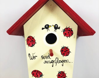 Może przedstawiać: Biały domek dla ptaków z czerwonym dachem i małym otworem, przez który ptaki mogą wejść. Domek jest ozdobiony biedronkami i napisem "Wir sind ausgeflogen..." czarnym kolorem.