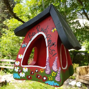 May include: A red wooden birdhouse with a black roof. The birdhouse is painted with flowers, butterflies, and the words "Bei Rudi, Pieps!"