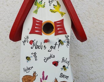 Puede incluir: Una casita para pájaros de madera pintada a mano con techo rojo y cuerpo blanco. La casita presenta pájaros, abejas y llamas pintados, junto con el texto "Mach's gut!" y varios nombres. La base tiene un paisaje pintado.