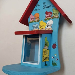 May include: A blue wooden house with a red roof and a window. The house is decorated with painted gnomes and the words "Auf neuen Zielen..." and "Colin Schmidle Kindergarten".
