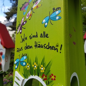 May include: A green wooden birdhouse with a white roof and a cutout for birds to enter. The birdhouse is decorated with colorful dragonflies and flowers. The text "Wir sind alle aus dem H&auml;uschen!" is painted on the front of the birdhouse.