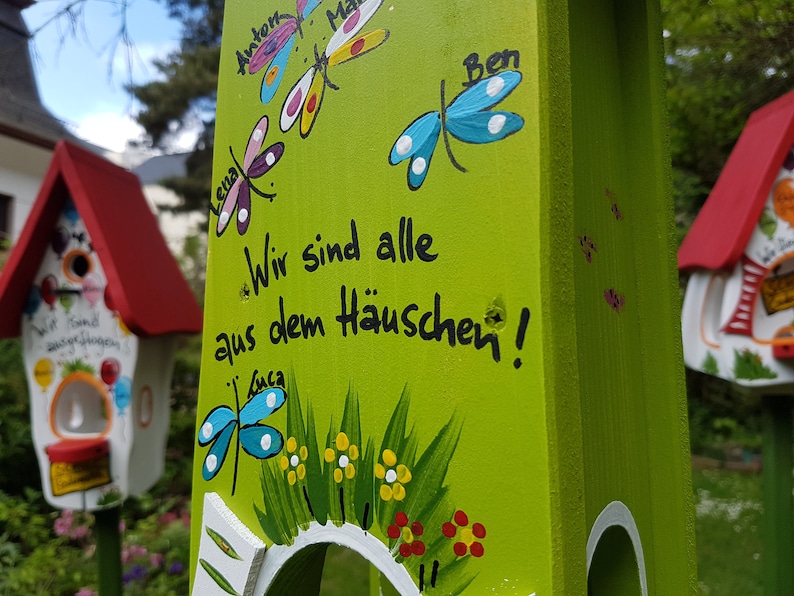 May include: A green wooden birdhouse with a white roof and a cutout for birds to enter. The birdhouse is decorated with colorful dragonflies and flowers. The text "Wir sind alle aus dem H&auml;uschen!" is painted on the front of the birdhouse.