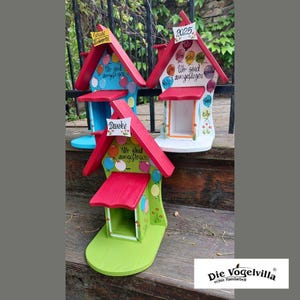 May include: Three brightly painted birdhouses with red roofs and various text. The birdhouses have blue, white, and green bases, decorated with balloon and flower designs. The text includes "Danke" and "Wir sind ausgeflogen."