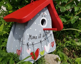 Puede incluir: Una casita de pájaros gris y roja con un patrón de lunares blancos y un techo rojo. La casita tiene una pequeña abertura para que los pájaros puedan entrar y un letrero que dice "Mama ist die Beste" con corazones colgando de una cuerda.