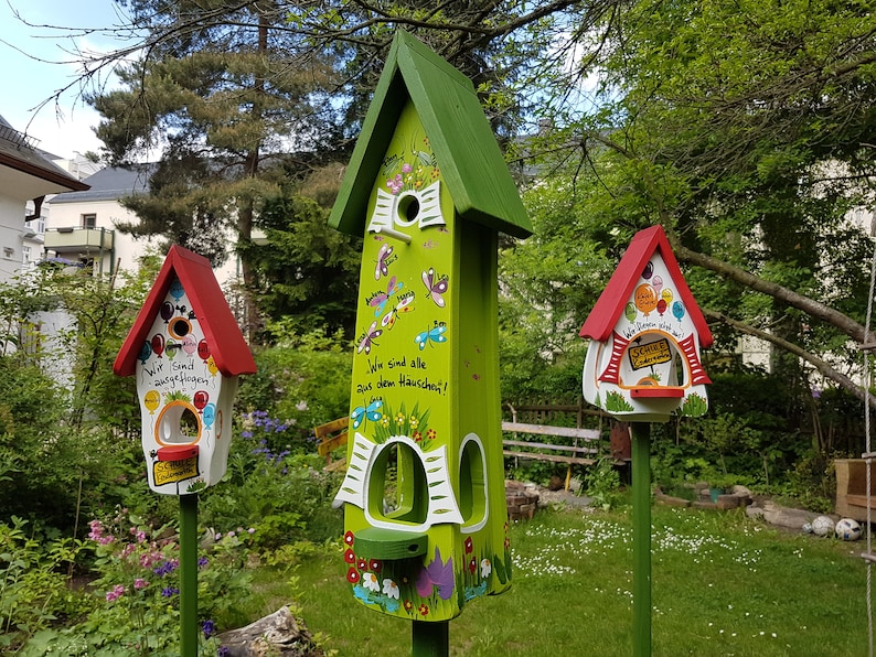 May include: Three colorful birdhouses with green, red, and white paint. The birdhouses have different designs and messages. The largest birdhouse is green with a white roof and has the text "Wir sind alle aus dem Hauschen!" The middle birdhouse is white with a red roof and has the text "Wir sind ausgeflogen!" The smallest birdhouse is white with a red roof and has the text "Schule ist cool!"