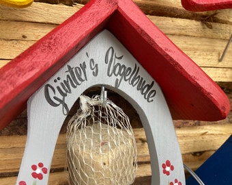 Pode incluir: Uma casa de pássaros vermelha e cinza com o texto "Günter's Vogelbude" pintado na frente. Um saco de rede cheio de sementes para pássaros pendura em um gancho na frente da casa de pássaros.