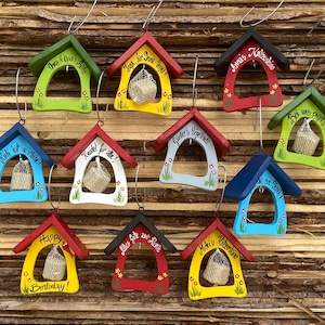 Op de afbeelding: Tien kleine houten vogelhuisjes met verschillende kleuren verf en boodschappen. Elk vogelhuisje heeft een klein netzakje met een witte vulling erin. De vogelhuisjes hangen aan een houten lattenwand.