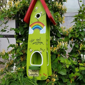 Op de afbeelding: Een groen en rood vogelhuisje met een regenboog geschilderd op de voorkant. Het vogelhuisje heeft een bord met de tekst "Schule Kindergarten" en heeft de namen van kinderen erop. Het vogelhuisje is omringd door groene bladeren.