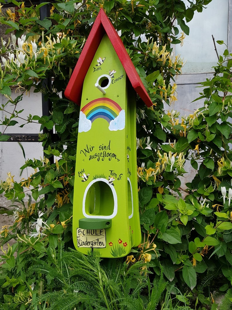 Op de afbeelding: Een groen en rood vogelhuisje met een regenboog geschilderd op de voorkant. Het vogelhuisje heeft een bord met de tekst "Schule Kindergarten" en heeft de namen van kinderen erop. Het vogelhuisje is omringd door groene bladeren.