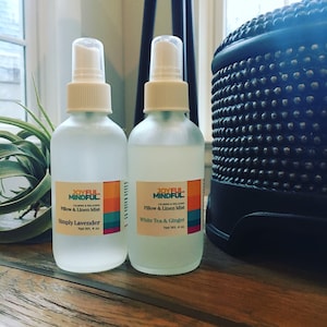 May include: Two glass spray bottles of Joyful Mindful Pillow & Linen Mist. One is labeled "Simply Lavender" and the other "White Tea & Ginger." Each bottle contains 113 g of product. The bottles have white spray tops and labels with orange, yellow, and blue accents.