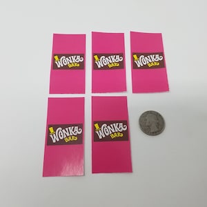 以下が含まれることがあります： 茶色、黄色、白のWonka Barのロゴが入った、ピンク色の長方形のWonka Barラベルが6枚。サイズの比較のために、ラベルの隣にコインがあります。ラベルは白い表面にあります。