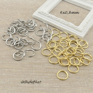 Könnte beinhalten: Nahaufnahme von zwei Stapeln Sprungringen. Ein Stapel ist silber und der andere ist gold. Die Sprungringe haben einen Durchmesser von 6 mm und eine Dicke von 0,8 mm. Der Text "6x0,8mm" ist im Bild sichtbar.