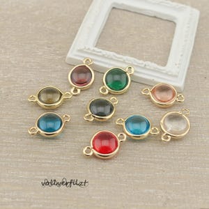 Könnte beinhalten: Eine Sammlung farbenfroher, runder Edelsteinanhänger mit goldfarbenen Metalleinfassungen. Die Edelsteine sind in verschiedenen Farben erhältlich, darunter Rot, Grün, Blau und Klar. Die Anhänger haben kleine Schlaufen zum Befestigen an Schmuck.