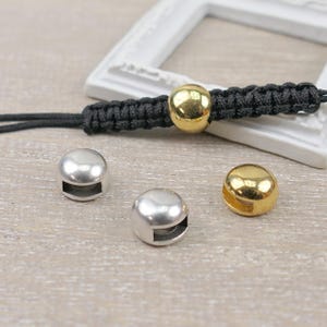 Könnte beinhalten: Ein schwarzes Kordelarmband mit einer einzelnen polierten goldfarbenen Kugelschmuckperle. Zwei weitere lose Kugeln, eine silberfarbene und eine goldfarbene, liegen daneben. Jede Perle hat eine rechteckige Öffnung an der Unterseite.